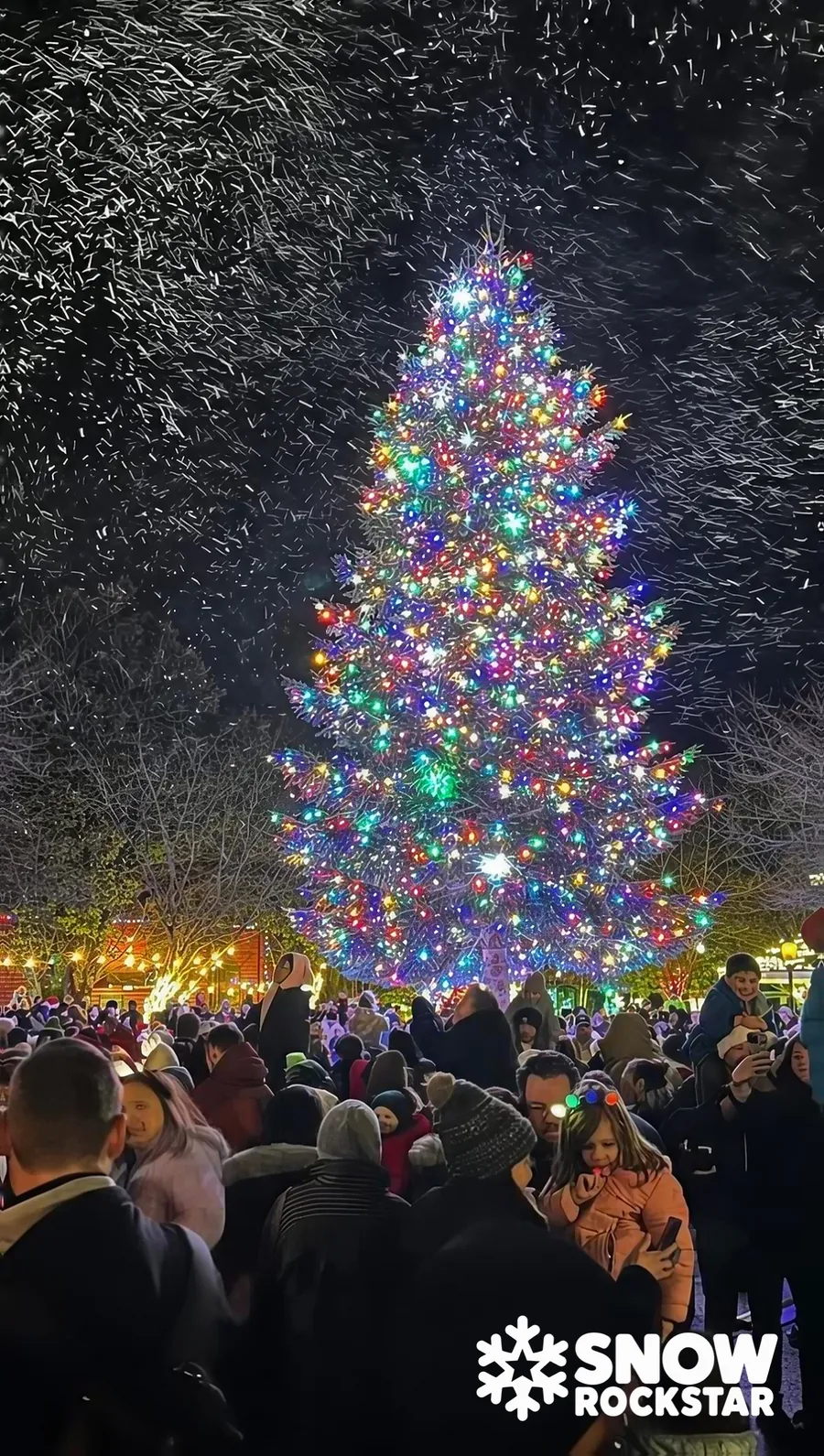 A huge multicolor-lit Christmas tree surrounded by a crowd as snow swirls through the air